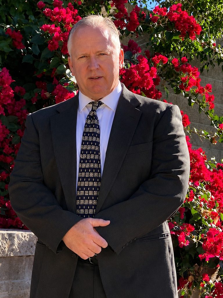 Private Investigator Phoenix, AZ in a sharp suit stands before red flowers, sunlight on his face.